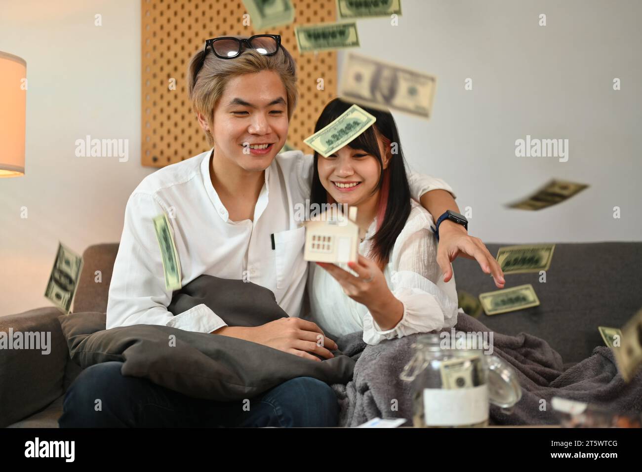 Glückliches, junges Paar, das Hausmodell hält, auf der Couch sitzt, mit Dollarscheine, die von oben fallen Stockfoto