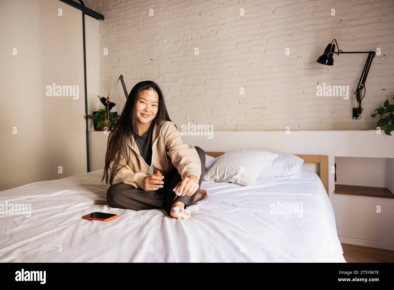 Junge chinesische, fröhliche Frau, die zu Hause Pediküre auf dem Bett macht Stockfoto