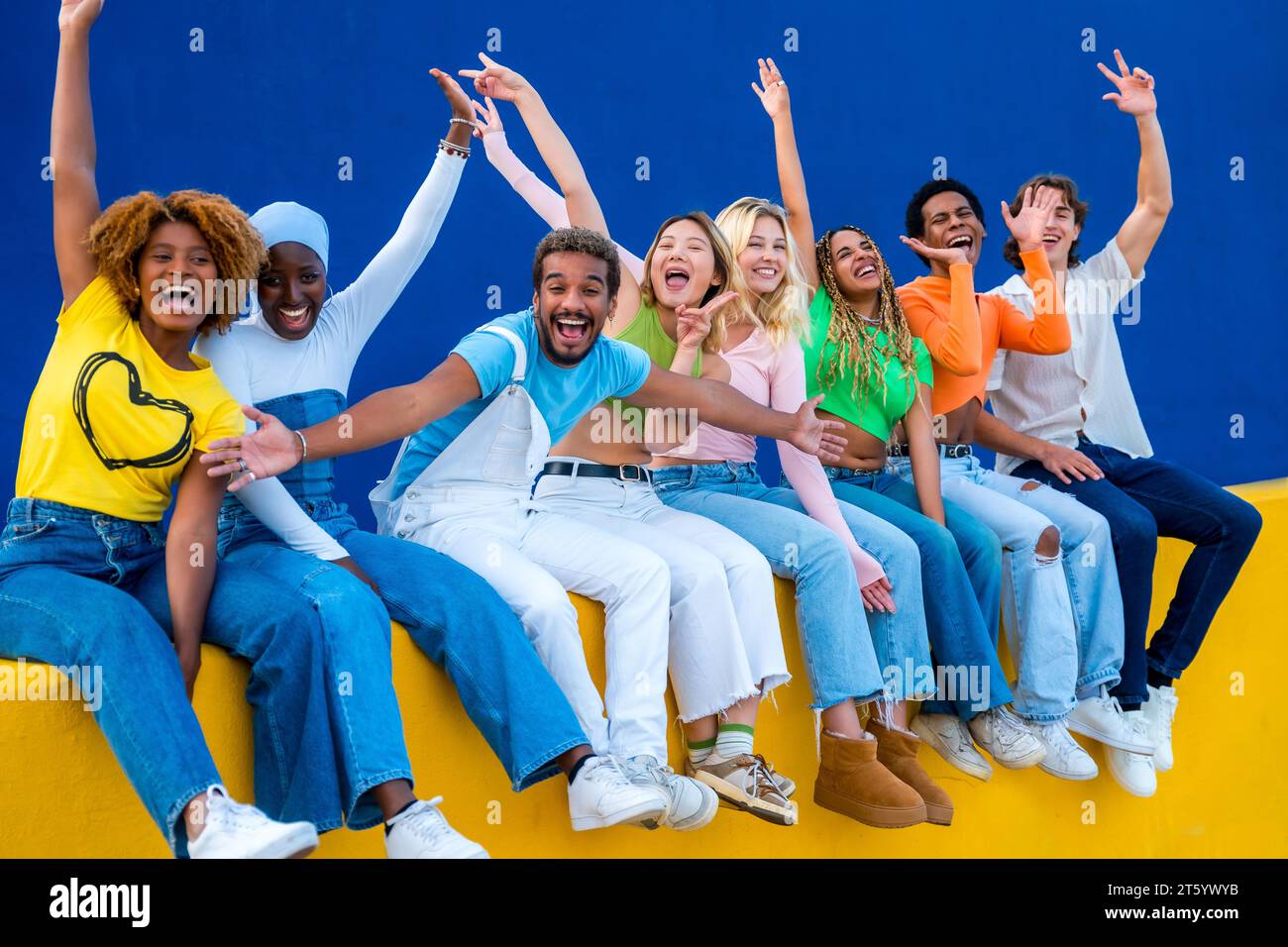 Fröhliche, multiethnische Freunde heben die Hände und lächeln, sitzen an einer Außenwand Stockfoto