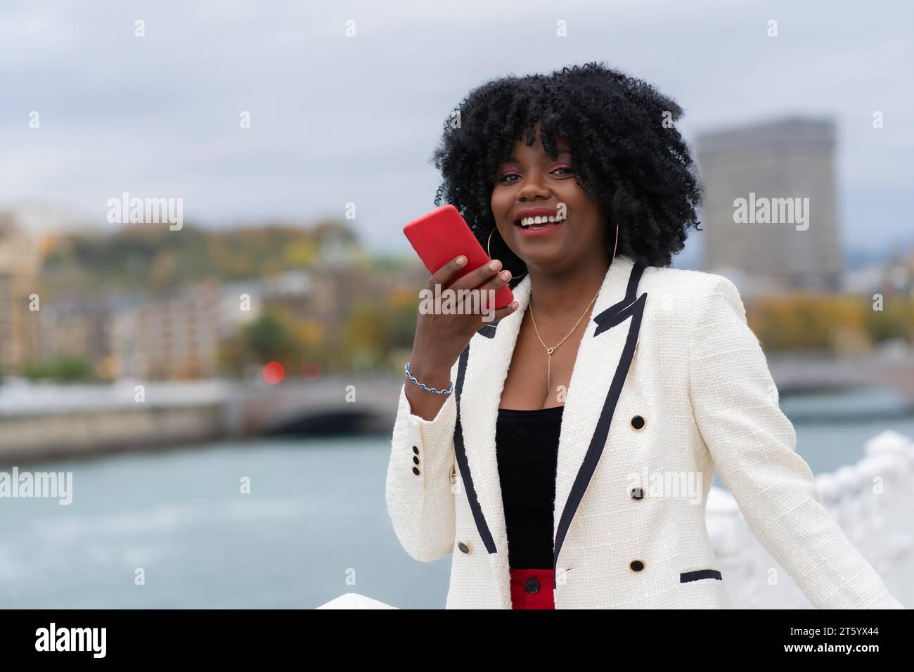 Elegante afroamerikanische Geschäftsfrau, die draußen neben einem städtischen Fluss eine Sprachnachricht schickt Stockfoto