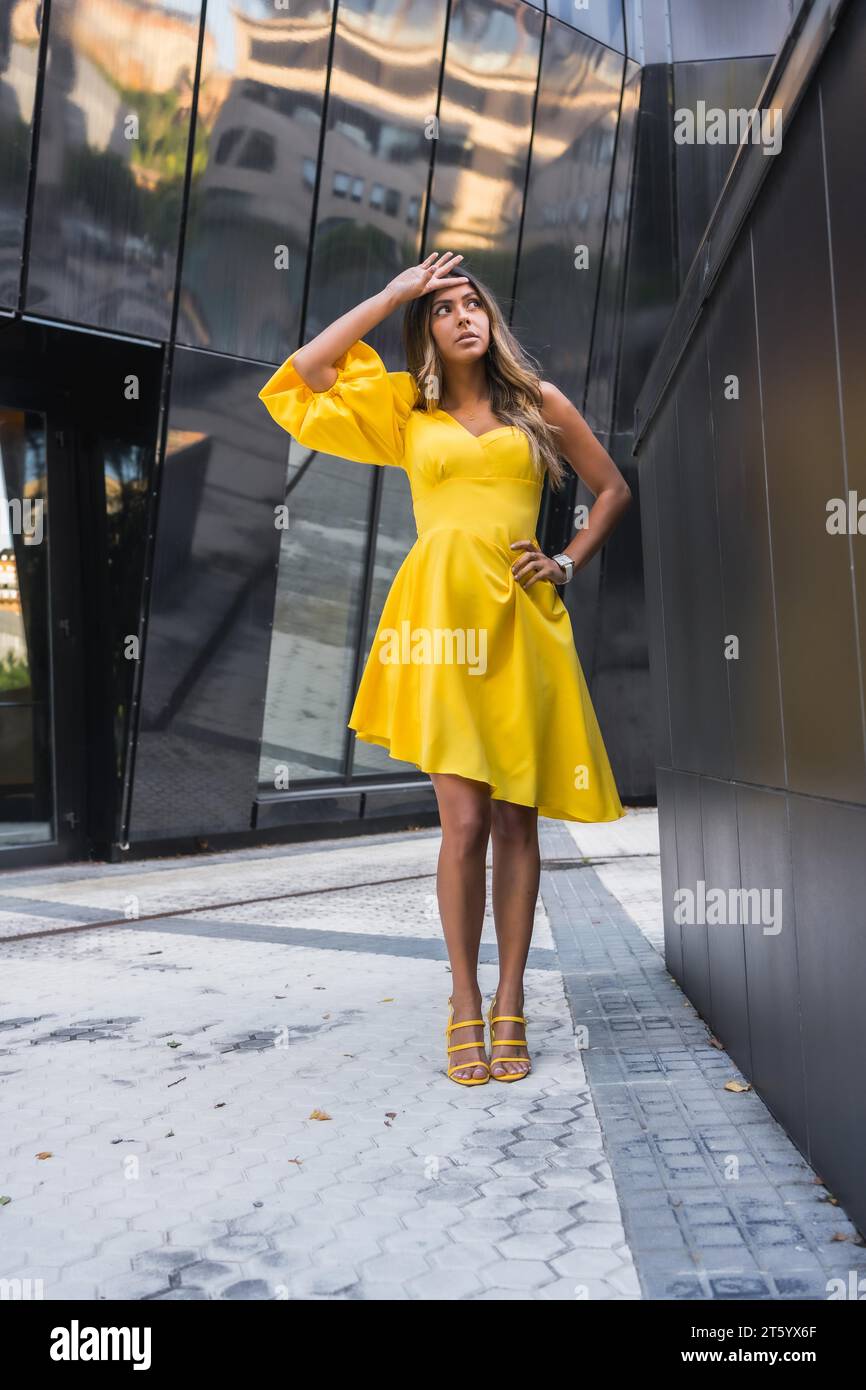 Vertikales Porträt eines schönen weiblichen Models mit gelbem Kleid, das in einer modernen Stadt posiert Stockfoto
