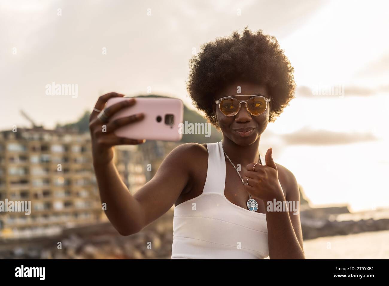 Afrikanische Frau, die sich bereit macht, ein Selfie zu machen, das draußen neben dem Meer steht, während Sonnenuntergang Stockfoto