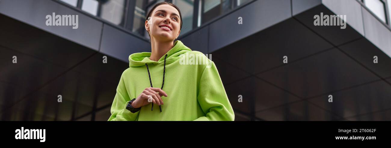 Niedriger Blickwinkel auf eine glückliche Sportlerin in einem übergroßen Hoodie, die draußen lächelnd ist, horizontales Banner Stockfoto