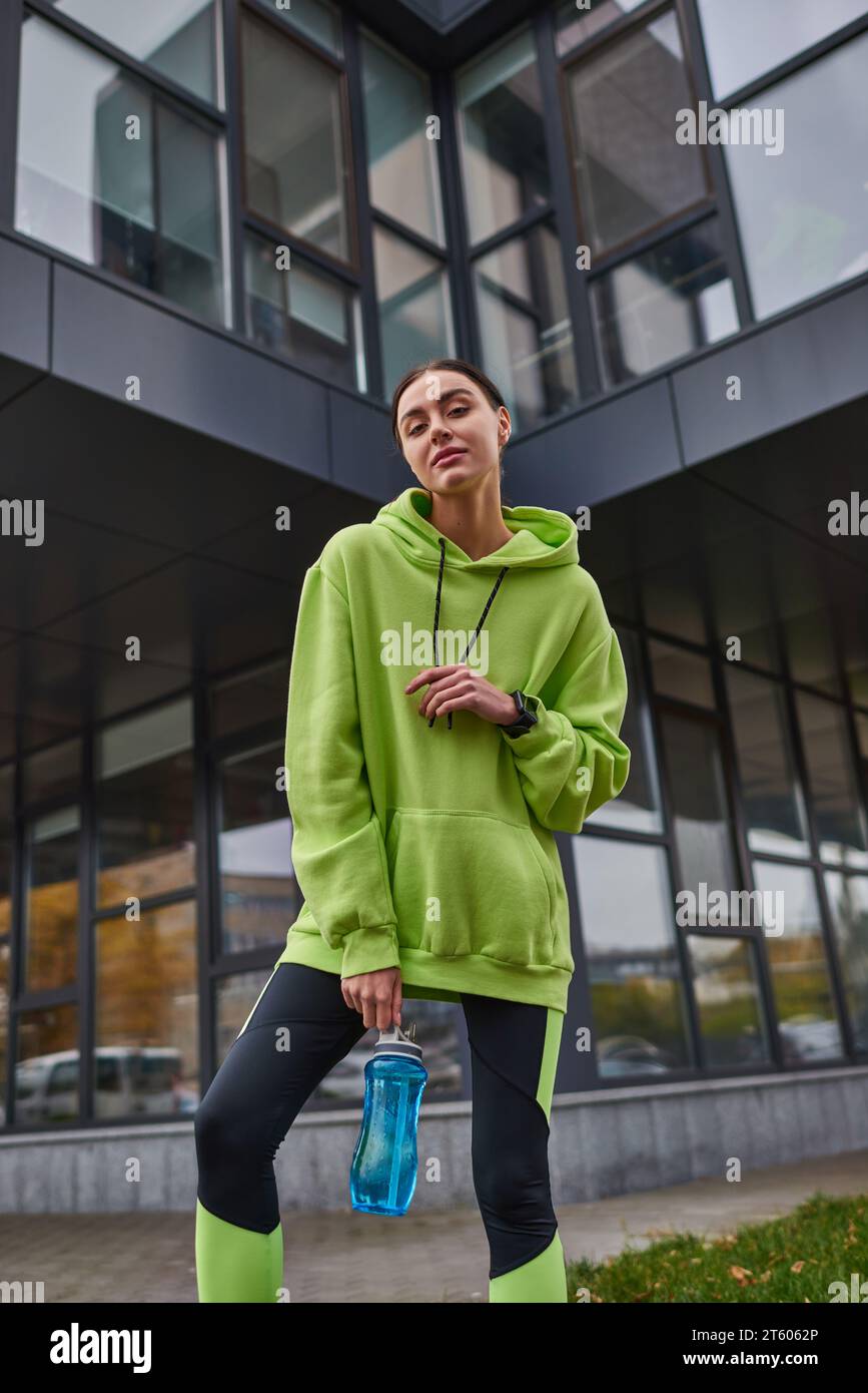 Niedriger Blickwinkel, Sportlerin in übergroßem Hoodie und Leggings mit Flasche und Wasser im Freien Stockfoto