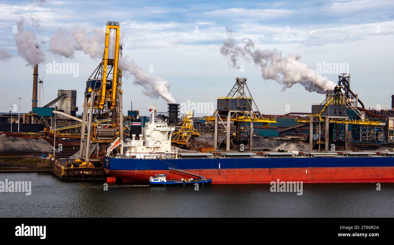 Frachtschiff, das in einem Industriehafen mit Kränen und Schornsteinen verladen wird, die Rauch gegen bewölkten Himmel ausstoßen Stockfoto