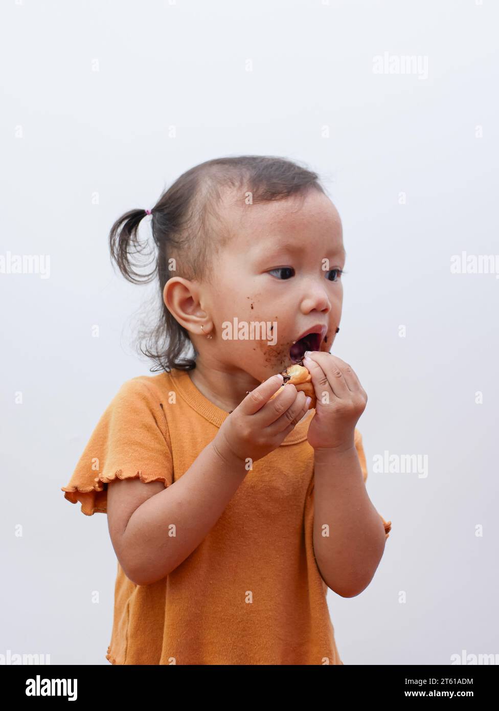 Süßes kleines asiatisches Mädchen, das süßes Donut oder Schokoladenbrot isst, Mund mit Schokolade bedeckt. Stockfoto