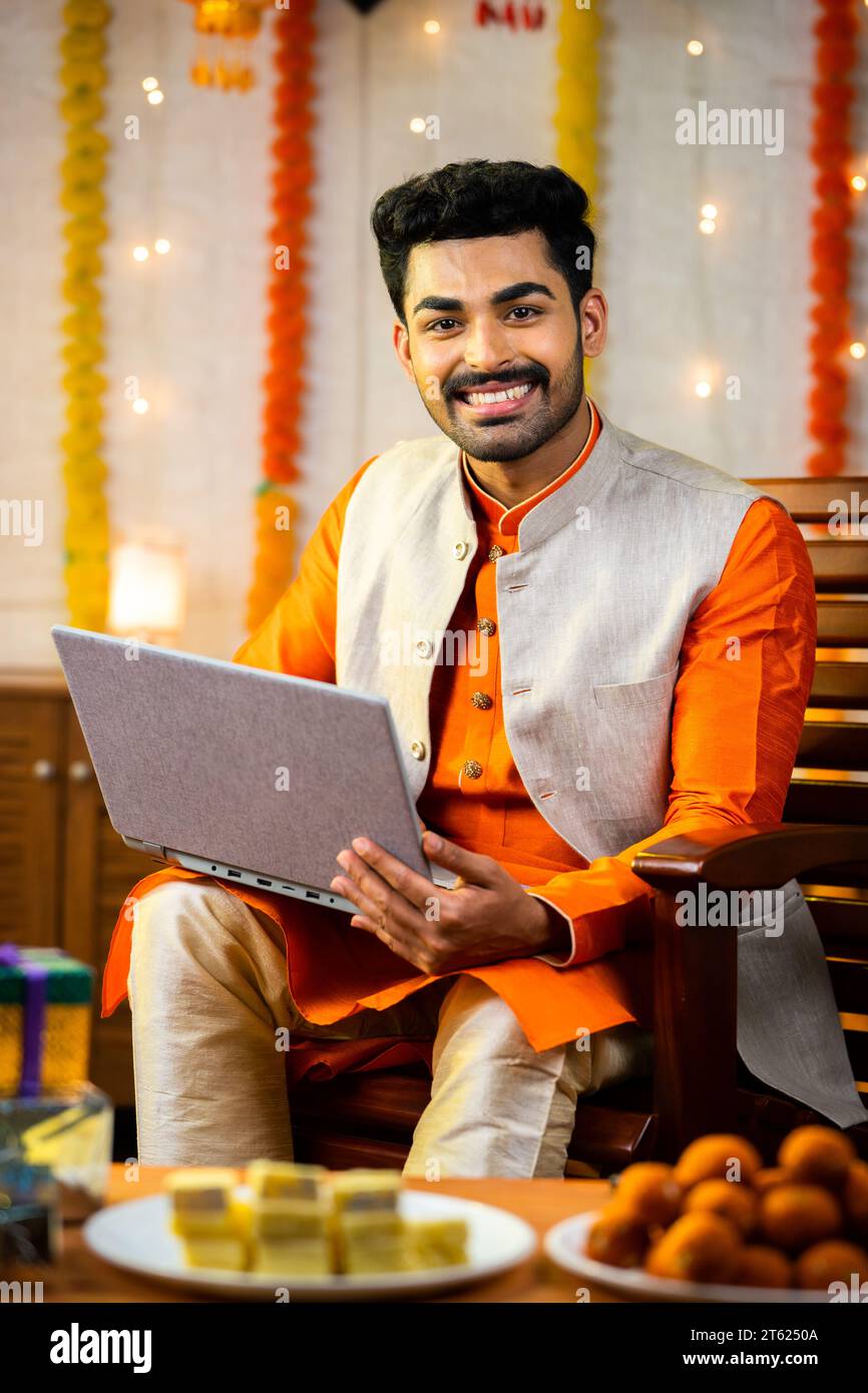 Vertikale Aufnahme eines glücklichen jungen indianers, der während des diwali Festivals Laptop benutzt, indem er zu Hause die Kamera betrachtet - Konzept der Festivalfeier, des Verkaufs und Stockfoto