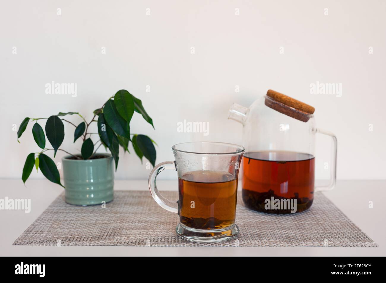 Schwarzer Tee in einer Glasschale und Glas-Teekanne auf einer Serviette und grüne Pflanze auf weißem Tisch mit gebläutem weißem Hintergrund Stockfoto