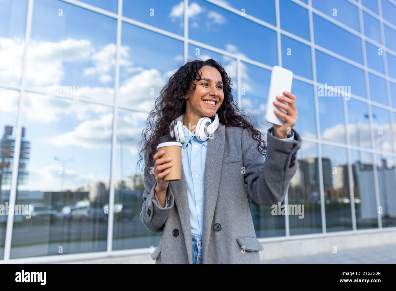 Junge schöne Frau, die in der Stadt vor einem Bürogebäude spaziert, hispanische Frau in einem Mantel mit Kopfhörern, eine Tasse heißes Getränk und ein Telefon, Musik hören und im Internet surfen. Stockfoto