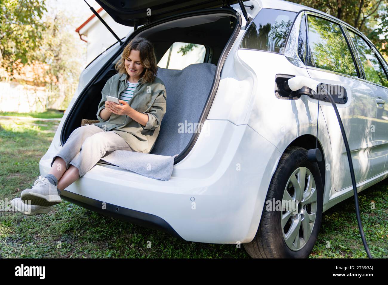 Frau mit Smartphone sitzt im Kofferraum eines Elektroautos. Stockfoto