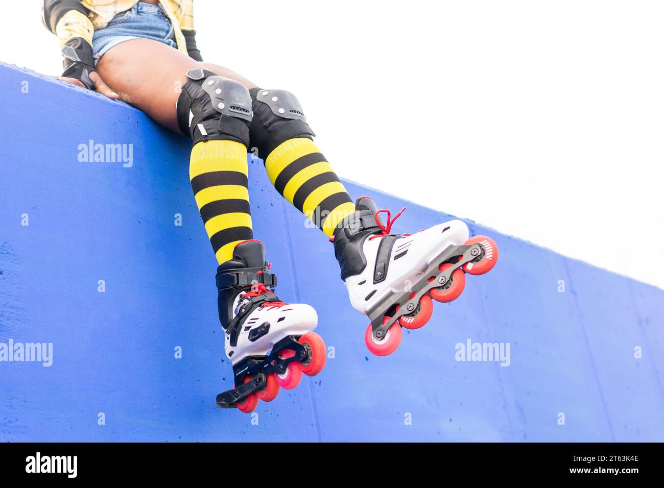 Niedriger Teil der afroamerikanischen Frau, die gelb gestreifte Socken und Rollschuhschuhe trägt, während sie am Skatepark vor klarem Himmel auf der blauen Wand sitzt Stockfoto