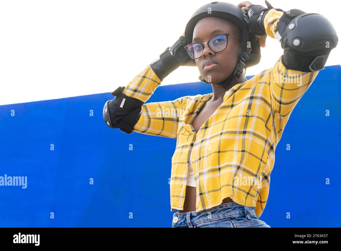 Nachdenkliche afroamerikanische junge Frau, die den Helm an der blauen Wand und dem klaren Himmel an sonnigen Tagen anpasst Stockfoto