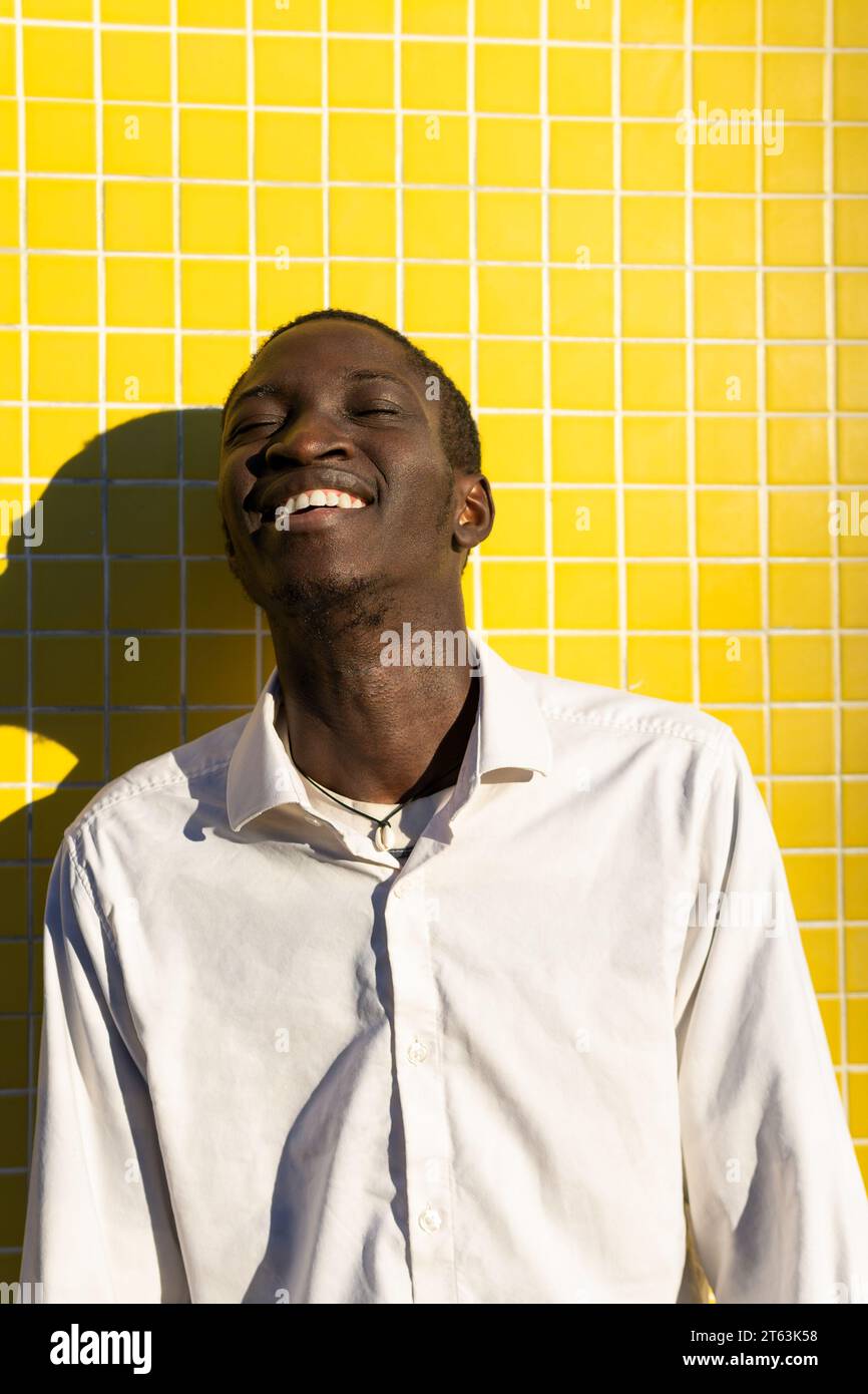 Fröhlicher schwarzer junger Mann lacht gegen eine leuchtend gelb gekachelte Wand, während die Sonne einen Schatten neben ihm wirft Stockfoto