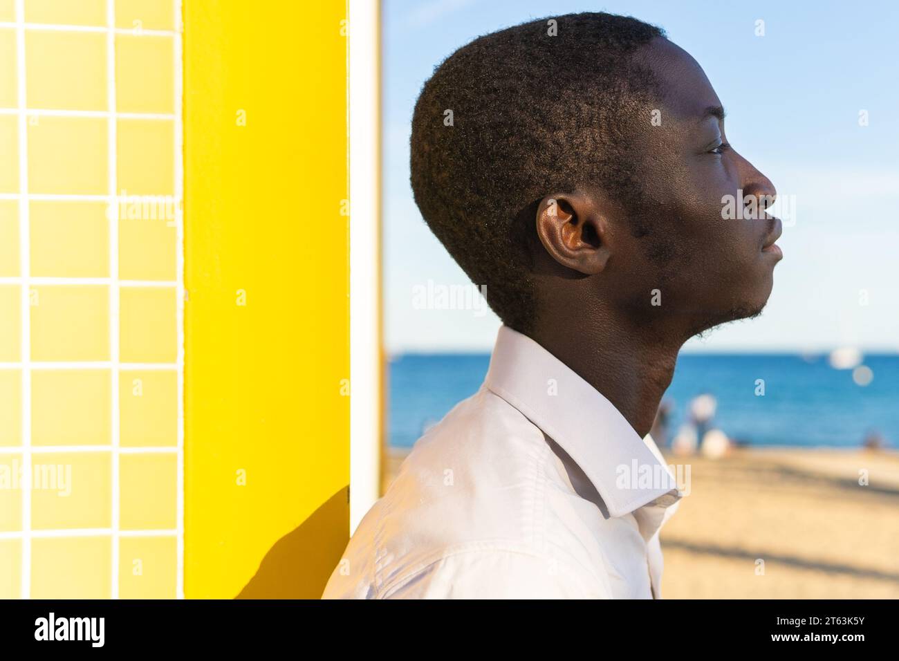 Ein junger afroamerikanischer Mann von der Seite blickt auf das Meer vor einem hellgelben Hintergrund und einer gefliesten Wand Stockfoto