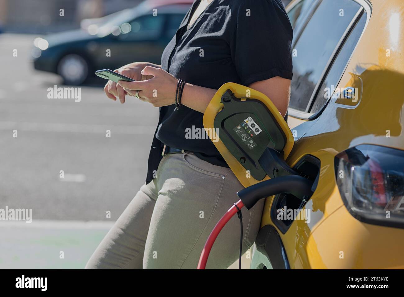 Eine Person in lässiger Kleidung steht neben einem gelben Auto und hält ein Smartphone, während ein Ladestecker für Elektrofahrzeuge an das Fahrzeug angeschlossen ist Stockfoto