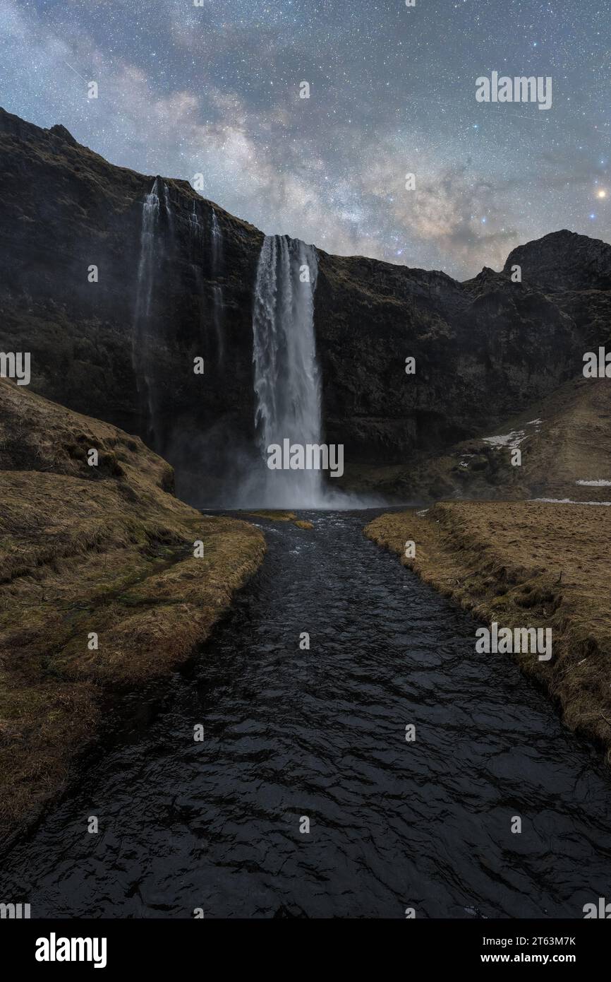 Eine majestätische Aufnahme von hohen Wasserfällen, die sich über eine dunkle felsige Klippe stürzen, mit einem ruhigen Fluss an der Basis, alles unter einem sternenklaren Nachthimmel in Island Stockfoto