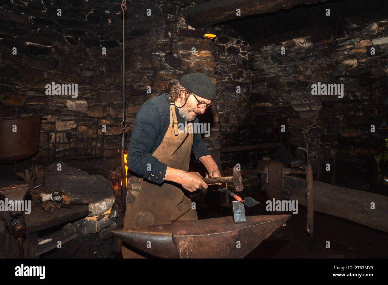 Seitenansicht des fokussierten Schmieds in traditioneller Kleidung schmiedet akribisch ein Metallstück in einer schwach beleuchteten Vintage-Werkstatt, umgeben von der alten Welt Stockfoto