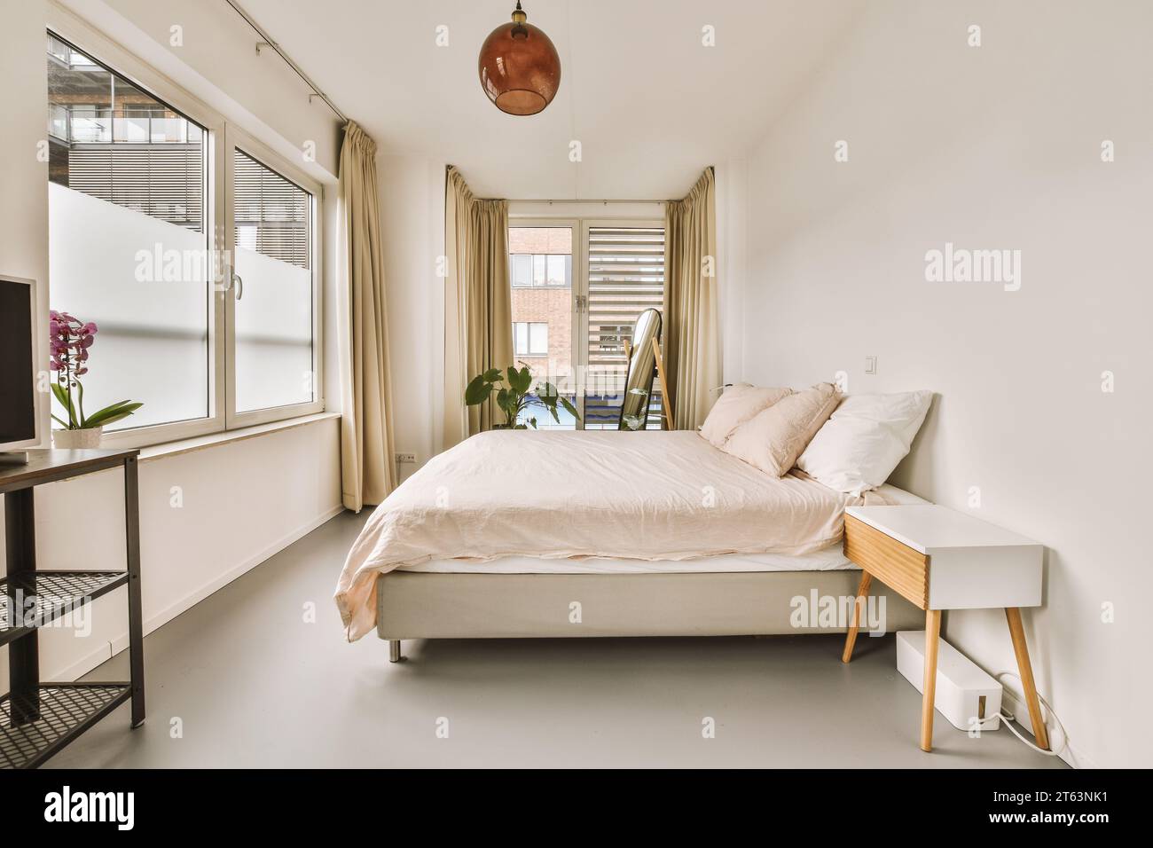 Das Innere des geräumigen weißen Schlafzimmers mit komfortablem Bett und Nachttisch vor dem großen Fenster in einem modernen Haus Stockfoto