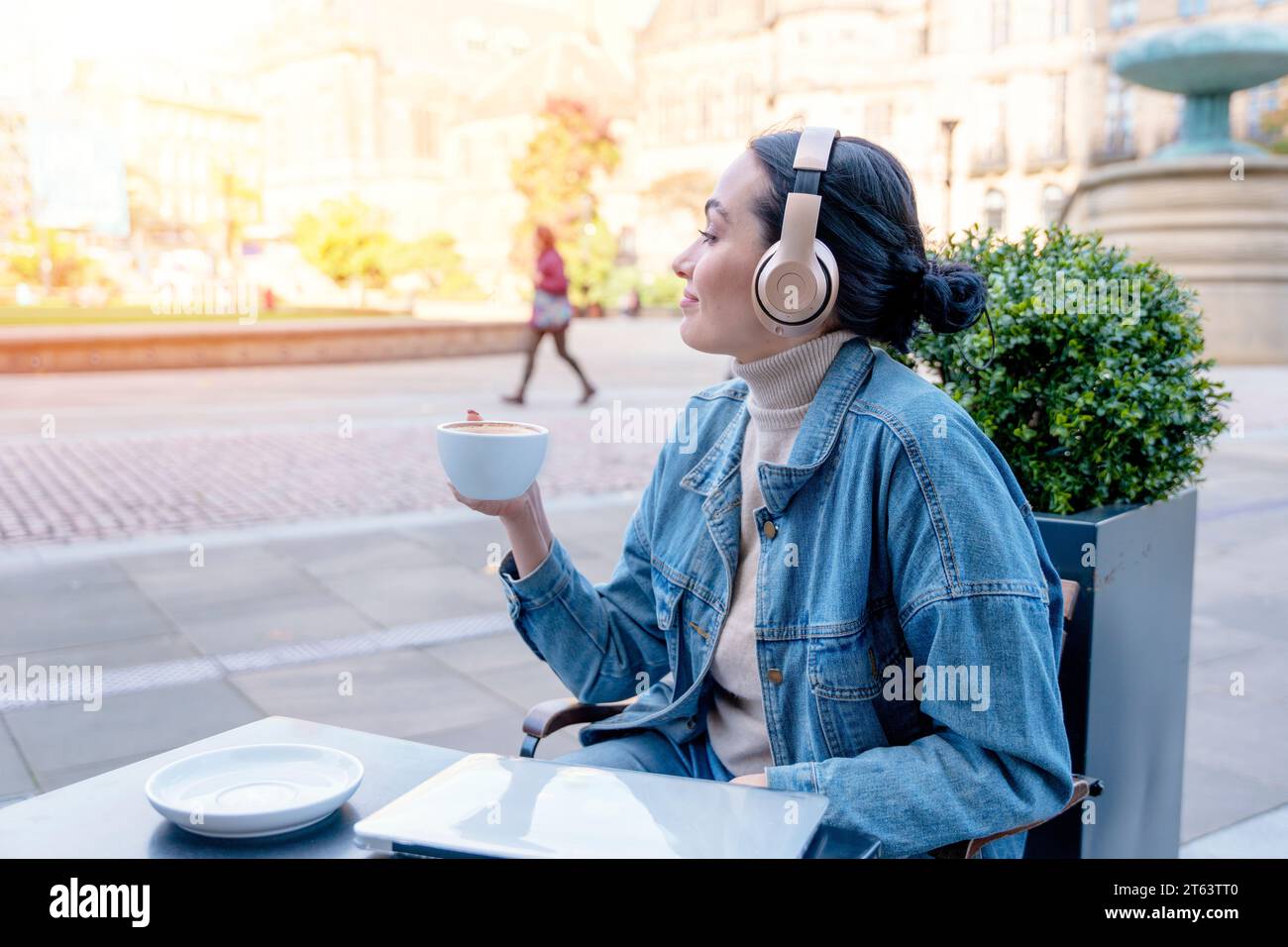 Frau in Jeansjacke und Kopfhörern trinkt Kaffee und hört Musik oder Hörbuch im Café. Lifestyle-Konzept Stockfoto