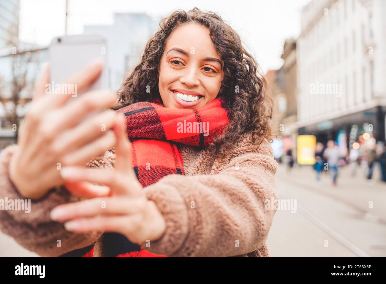 Eine fröhliche Frau, die Selfies mit einem Smartphone macht und telefoniert. Eine lockige braune Dame in einer Jacke und einem orangefarbenen Schal, die lächelt und in der EU reist Stockfoto