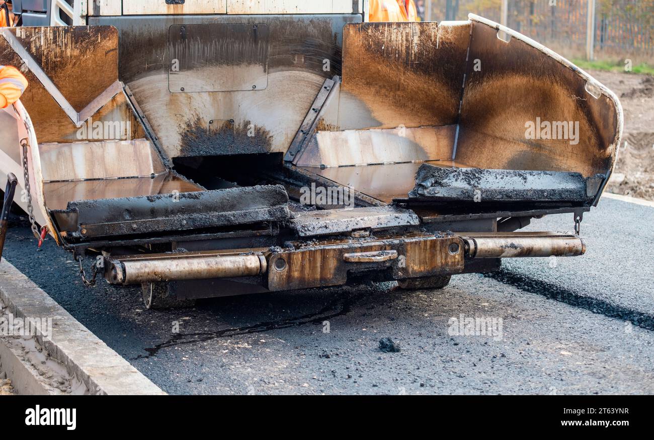 Asphaltfertiger mit heißem Asphalt, der neue Straßenoberflächen auf neuen Wohnbaustellen verlegt, und Straßenarbeiter in orangefarbenem Hi-viz als nächstes Stockfoto
