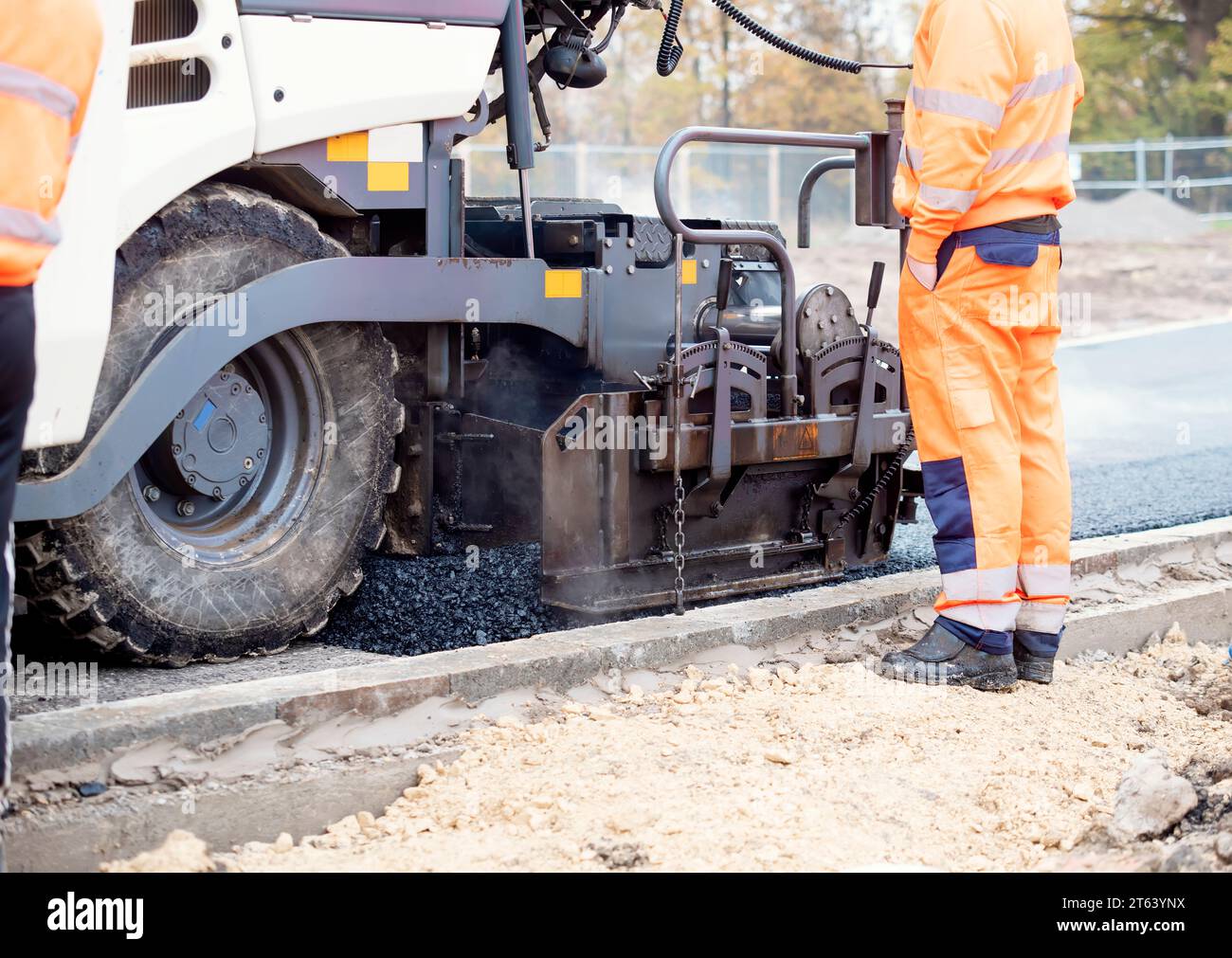 Asphaltfertiger mit heißem Asphalt, der neue Straßenoberflächen auf neuen Wohnbaustellen verlegt, und Straßenarbeiter in orangefarbenem Hi-viz als nächstes Stockfoto