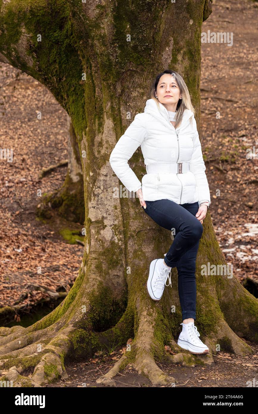 Frau, die auf einem moosbedeckten Baumstamm im Wald sitzt, eine weiße Jacke und eine schwarze Hose trägt Stockfoto