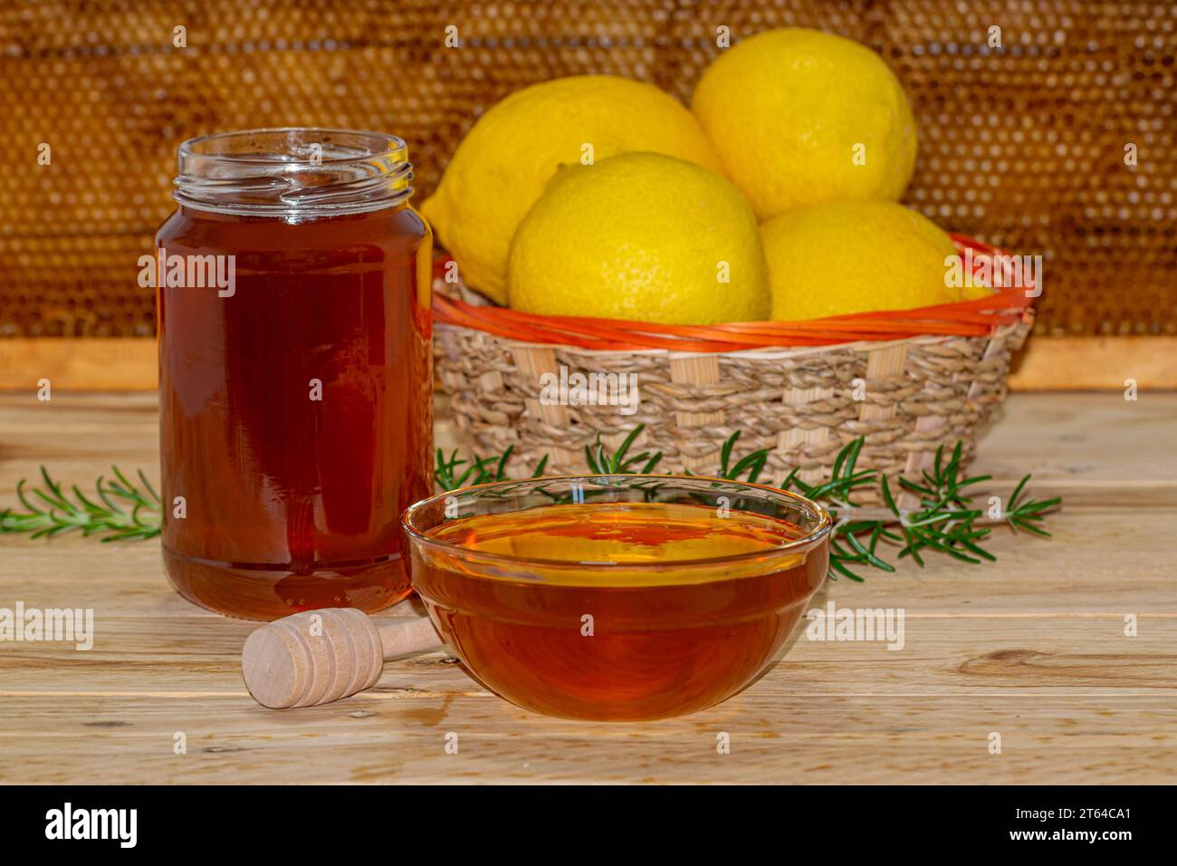 Honigglas ohne Deckel, Spender, eine Schüssel Honig, eine Rosmarinfeder und ein Korb Zitronen auf Holz mit einer echten Wabe im Hintergrund Stockfoto
