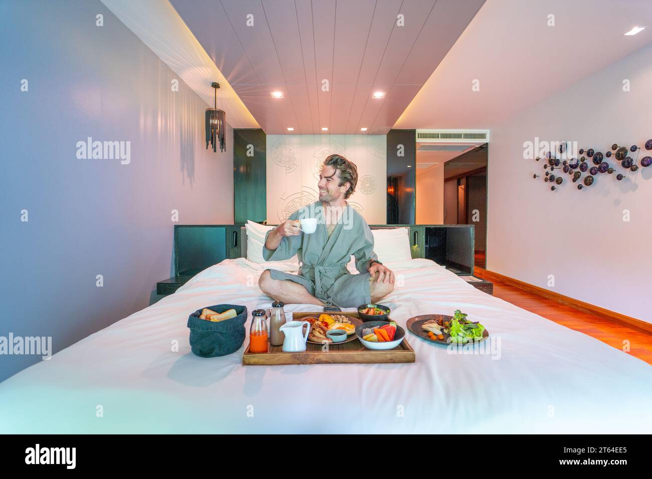 Gutaussehender Mann isst ein großes Zimmerservice-Frühstück im Komfort seines Hotelbetts Stockfoto
