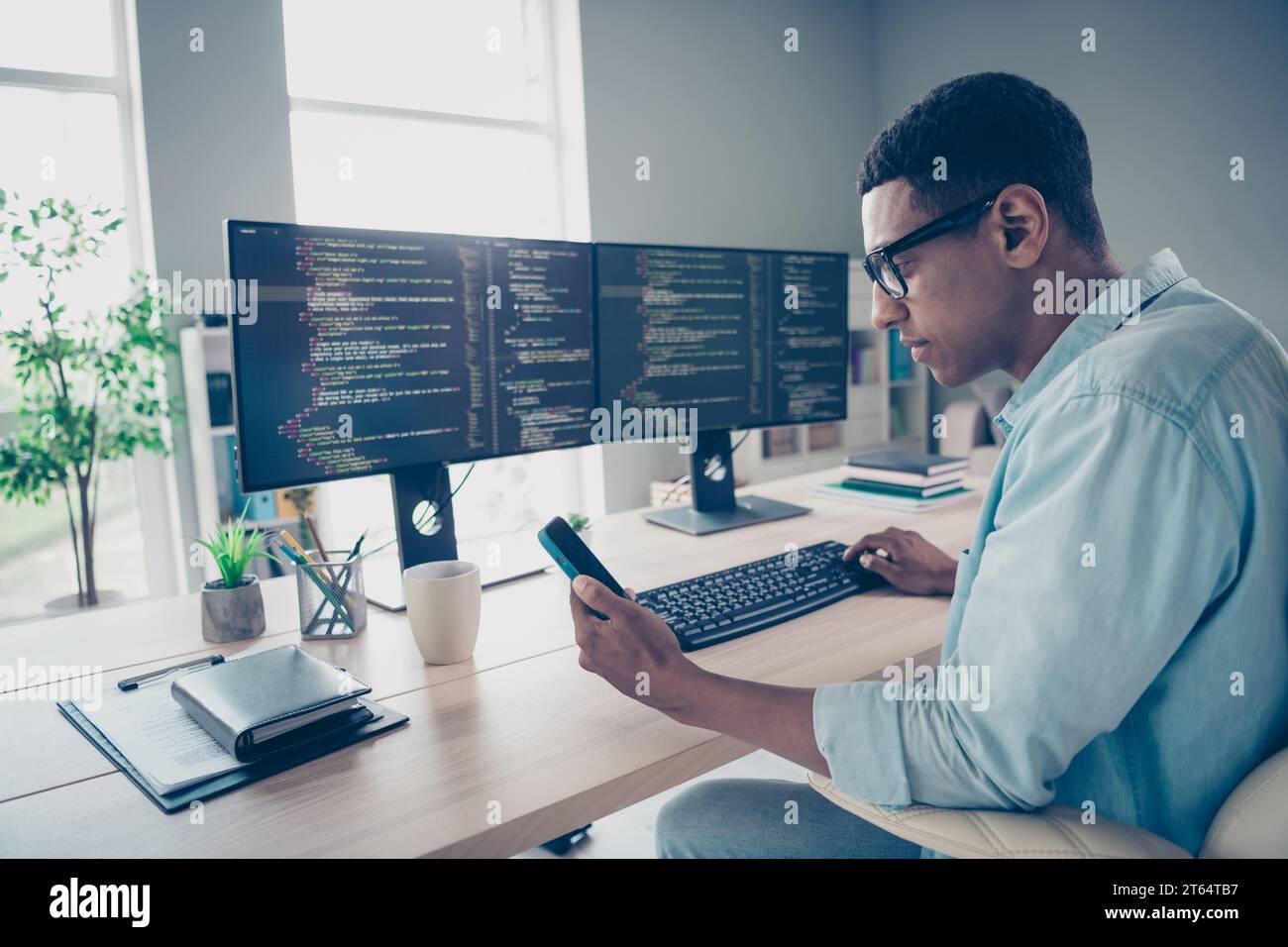 Foto eines klugen jungen Mannes tragen blaues, informelles Hemd, das kodiert, Computer-Bearbeitung, Datenbankcode-Programm, das Telefon im Werkstattbüro schreibt Stockfoto