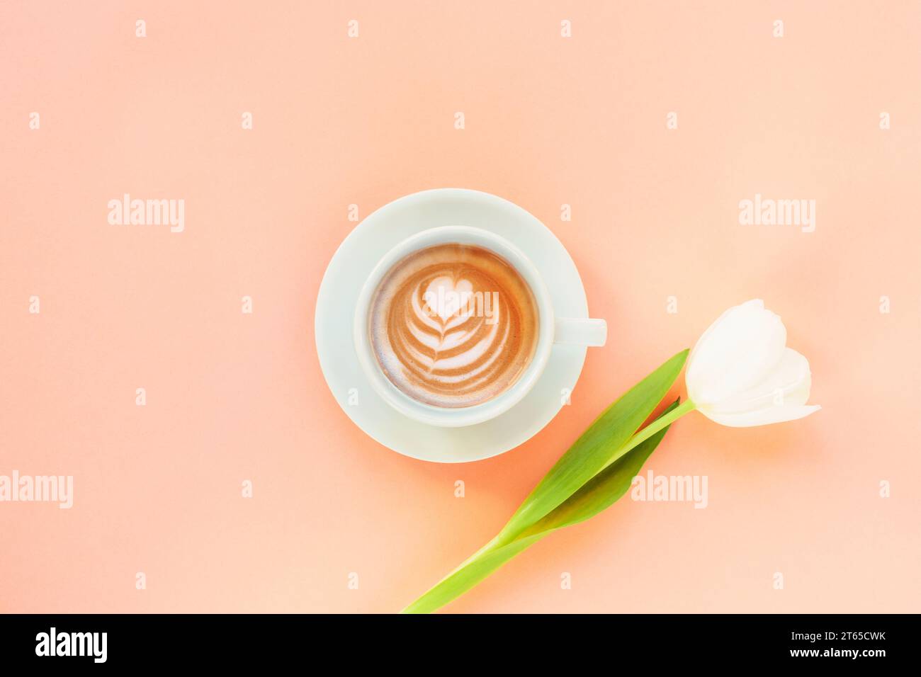 Tasse Kaffee mit Kaffeekunst und weißer Tulpe auf pfirsichfarbenem Hintergrund. Frauentag oder Muttertagskonzept. Draufsicht, flach, Kopierbereich. Stockfoto