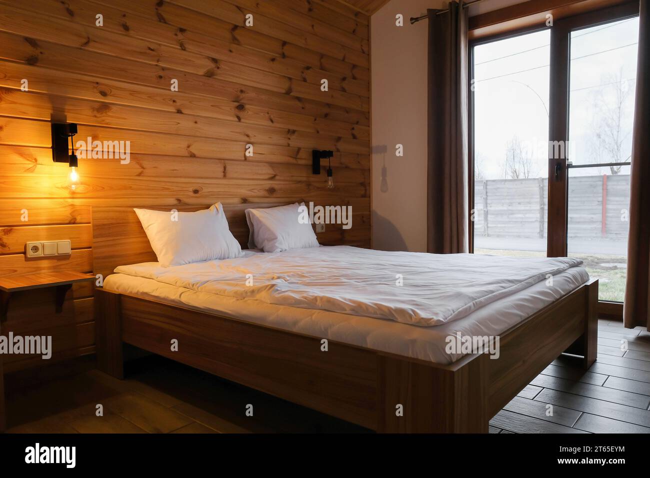 Schlafzimmereinrichtung in einem hölzernen Landhaus. Rustikale Blockhütte Schlafzimmer innen, gemütliches Bett durch das große Fenster Stockfoto