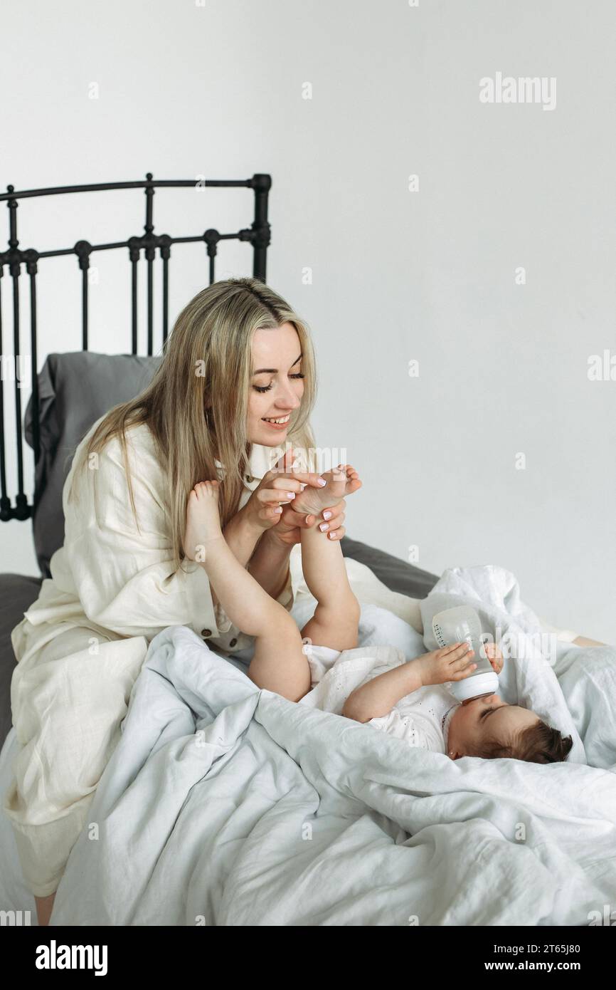 Mom kitzelt dem Kind die Füße, das Mädchen liegt auf dem Bett und trinkt Wasser aus einer Flasche. Vertikaler Rahmen. Stockfoto