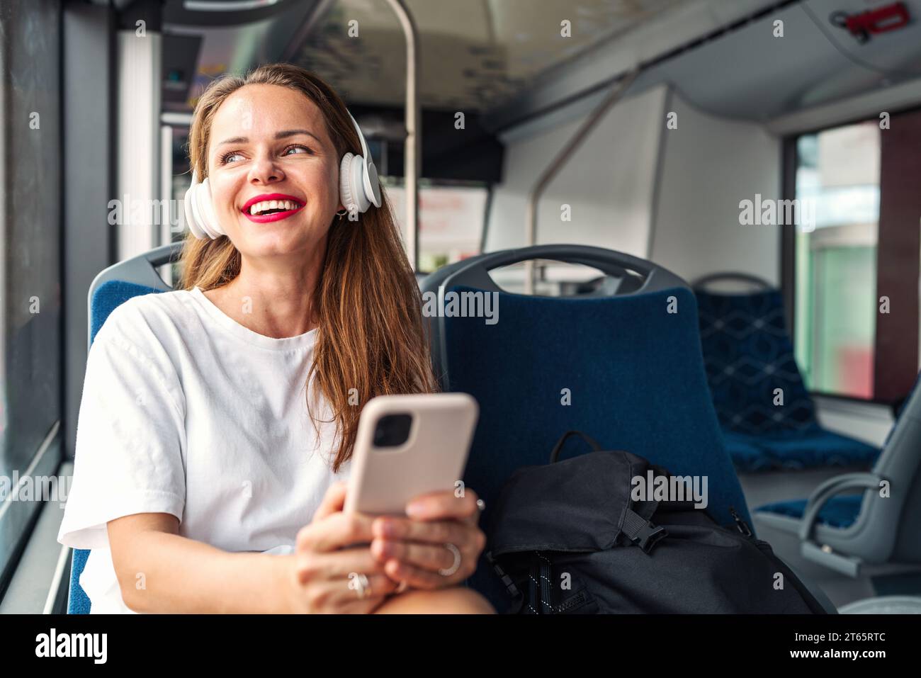 Glückliche Frau in Kopfhörern, die ihr Handy in der Hand hält und Musik hört, während sie mit dem Shuttlebus durch die Stadt fährt. Stockfoto