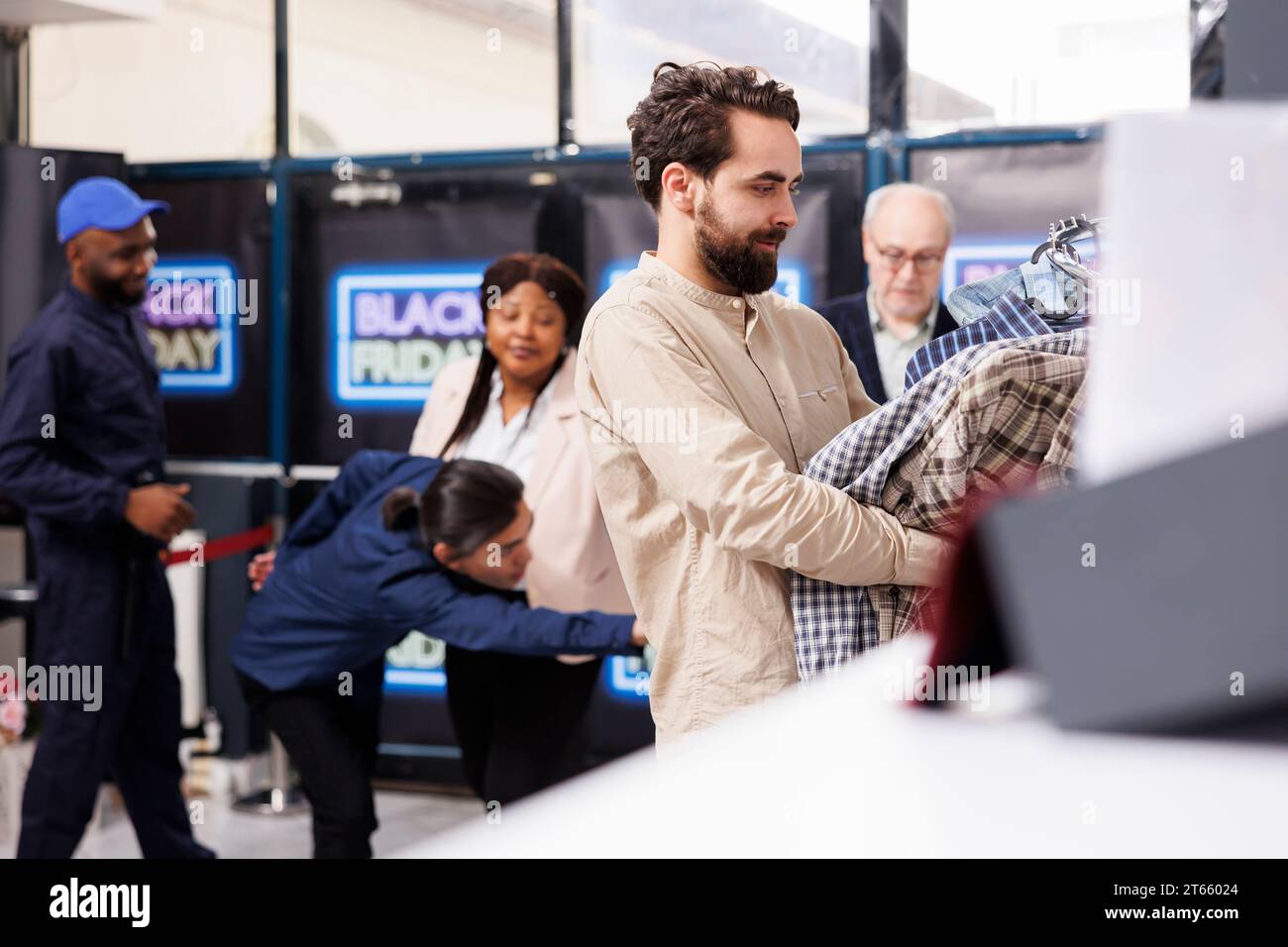 Junger Mann, der Kleidung in Händen hält, während er an der Kasse des Bekleidungsgeschäfts steht, Leute zahlen für Black Friday-Einkäufe. Männlicher Kunde genießt Rabatteinkäufe während des Verkaufs Stockfoto