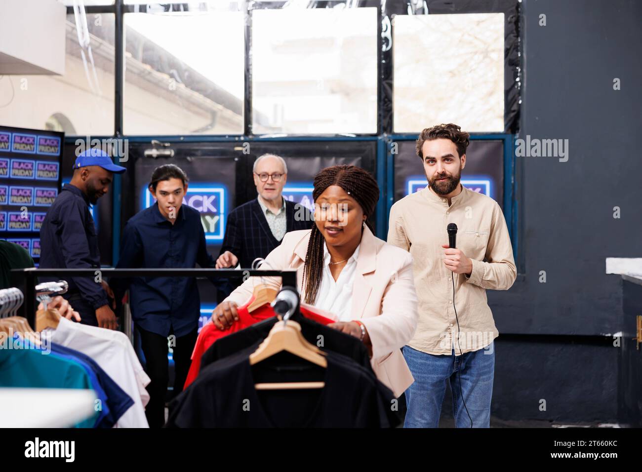 Journalist hält ein Mikrofon und blickt in die Kamera, während er über Black Friday-Verkäufe aus dem Einkaufszentrum berichtet. Eine Menge aufgeregter Leute, die an TV-Reportern vorbeilaufen, um die besten Angebote zu erhalten Stockfoto