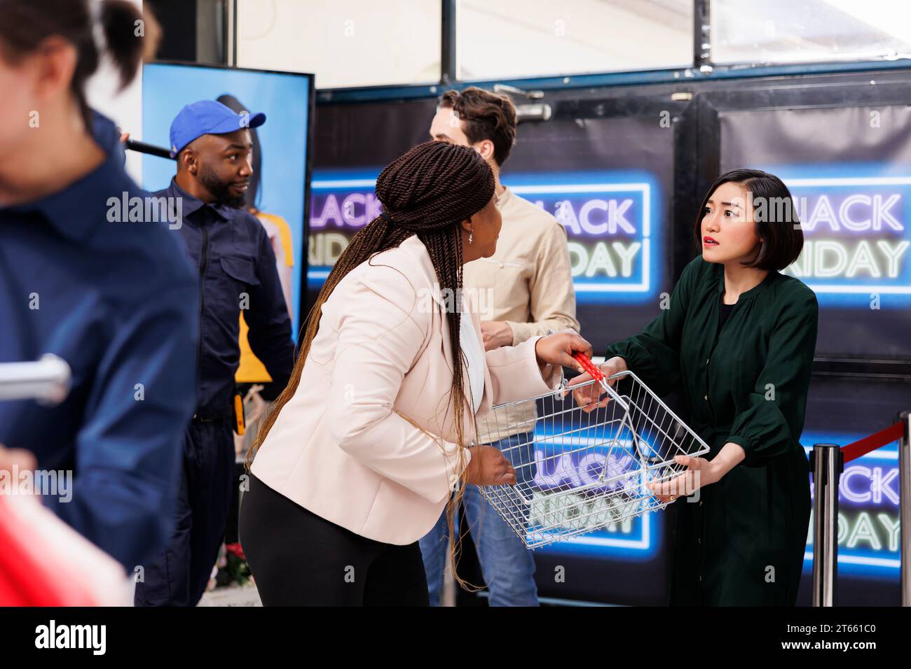 Während des Black Friday kämpfen die Leute dafür, Dinge zu kaufen. Zwei gierige, wütende, vielfältige Käuferinnen stehen am Eingang des Einkaufszentrums und kämpfen um den letzten Warenkorb, während sie nach Schnäppchen suchen Stockfoto