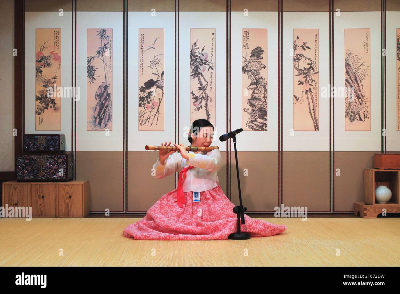 Eine Frau in historischen Kleidern spielt eine traditionelle koreanische Bambusflöte vor mehreren asiatischen Pinselgemälden an der Wand dahinter. Stockfoto