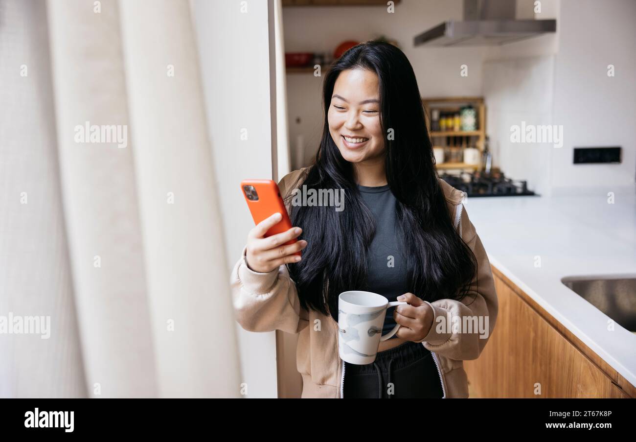 Junge, schöne Frau mit einem Becher und einem roten Telefon neben einem Fenster in einer Küche. Lächelnde Frau, die zu Hause eine Kaffeepause genießt. Stockfoto