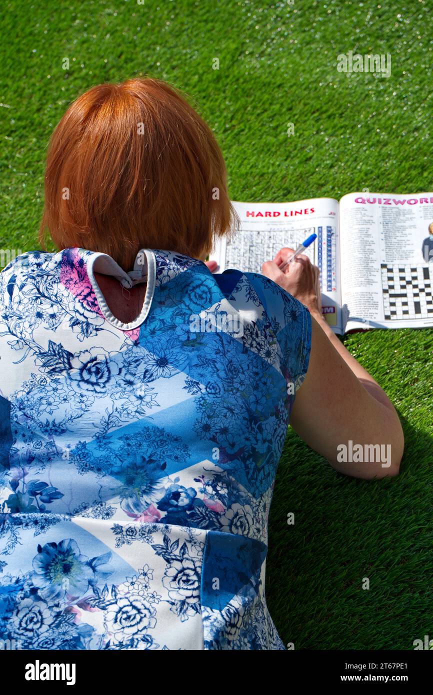 Frau mit rotem/Ingwerhaar in einem blau-rosa gemusterten Kleid, die auf ihrer Vorderseite auf Kunstrasen liegt, in der Sonne und macht ein Puzzlesuch Stockfoto