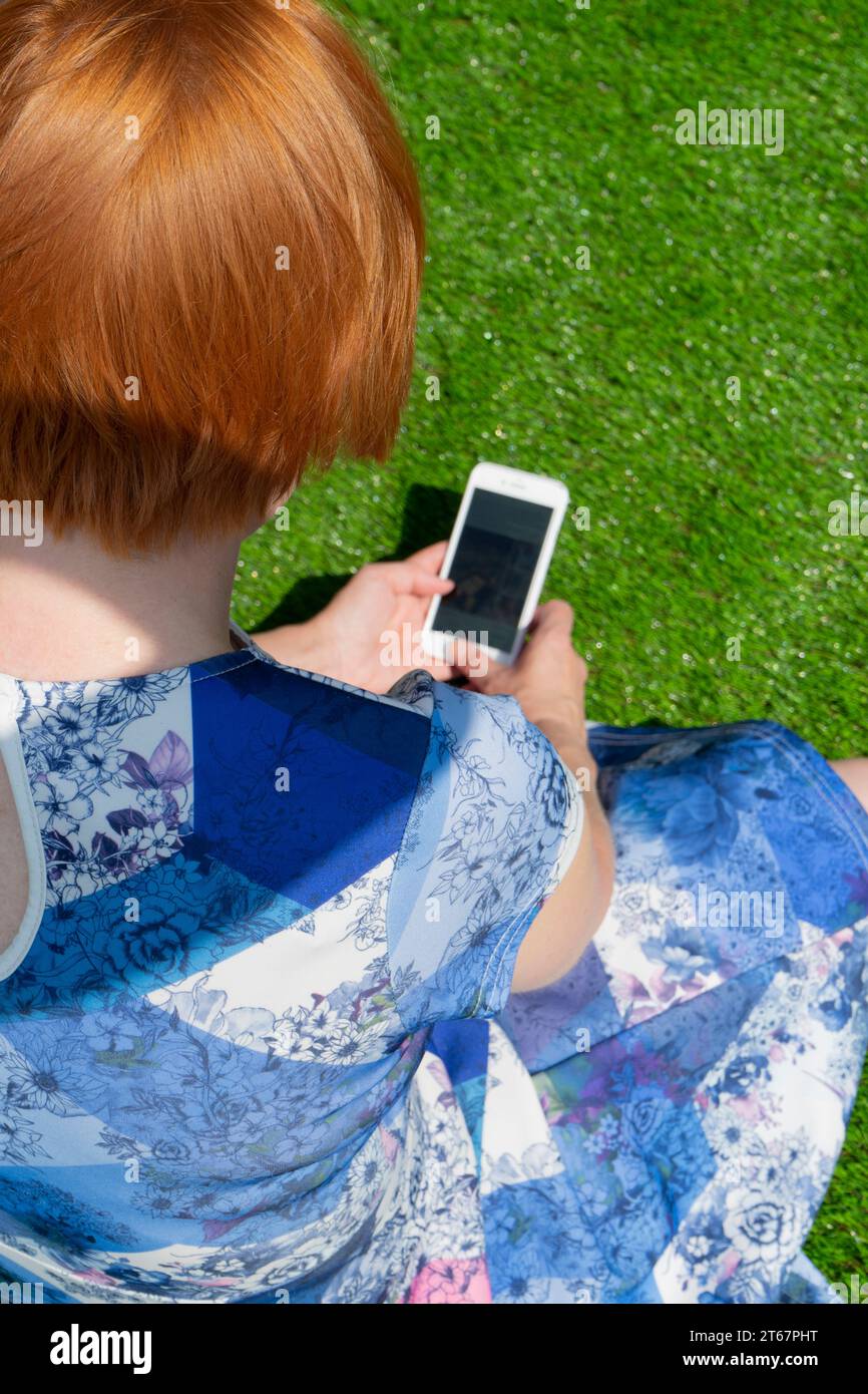 Eine rothaarige Frau in einem blau gemusterten Kleid, sitzend auf Kunstrasen in der Sonne, schaute hinunter und benutzte ihr iPhone Stockfoto