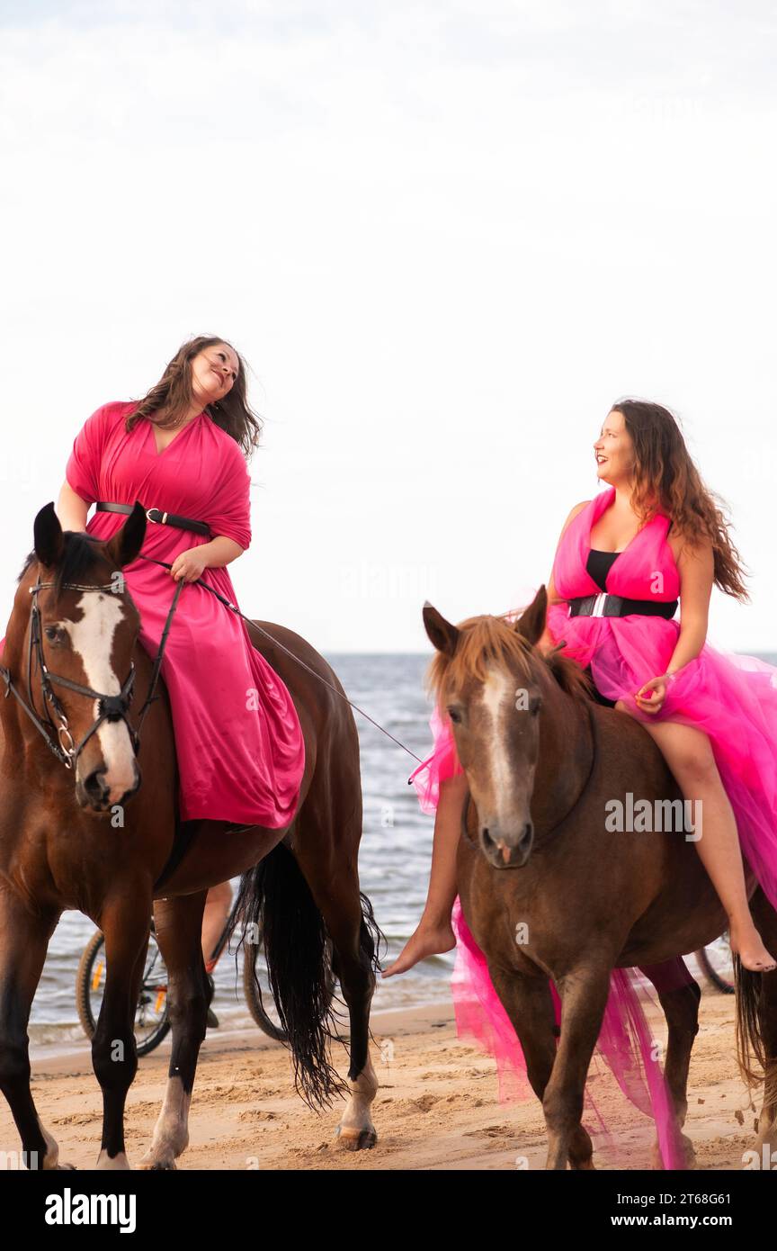 Zwei weibliche Reiterinnen in eleganten rosa Kleidern galoppieren bei Sonnenuntergang an einem malerischen Strand entlang, der sich am goldenen Himmel abhebt Stockfoto