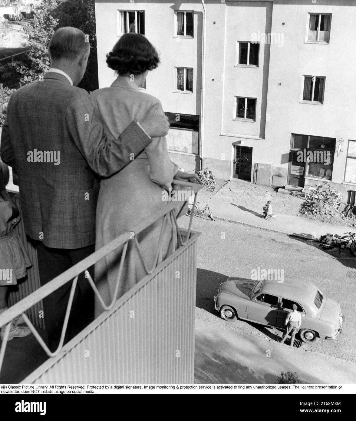 In den 1950er Jahren Ein Paar, das auf dem Balkon ihrer Wohnung auf die Straße unten blickt, wo ihr Auto geparkt ist und ein Kind auf einem Dreirad auf dem Bürgersteig fährt. Schweden 1955. Conard Stockfoto