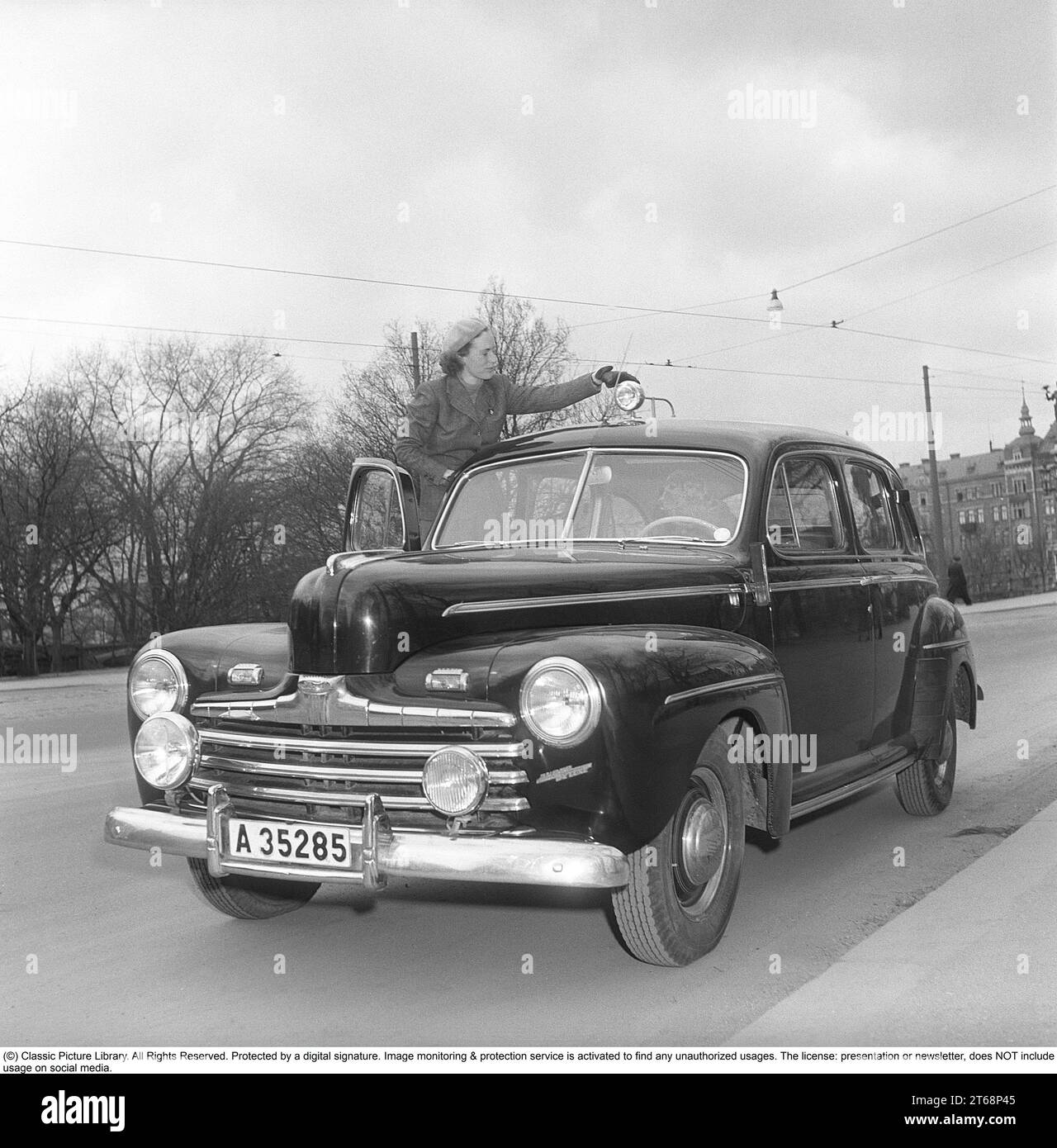 In den 1940er Jahren Eine junge Frau bei ihrem Pkw der amerikanischen Automarke Ford. Sie trägt einen zweiteiligen Anzug, einen Rock und eine kurze Jacke aus Fischgrätwolle. Sie steht auf den Stufen des Autos, um zum Dach zu gelangen, wo ein Suchscheinwerfer montiert ist. Manchmal muss sie angepasst werden. Ihr Hund kann am Lenkrad auf dem Fahrersitz sitzen. Das Foto wurde 1949 in Stockholm, Schweden, aufgenommen. Kristoffersson Ref. AY83-6 Stockfoto