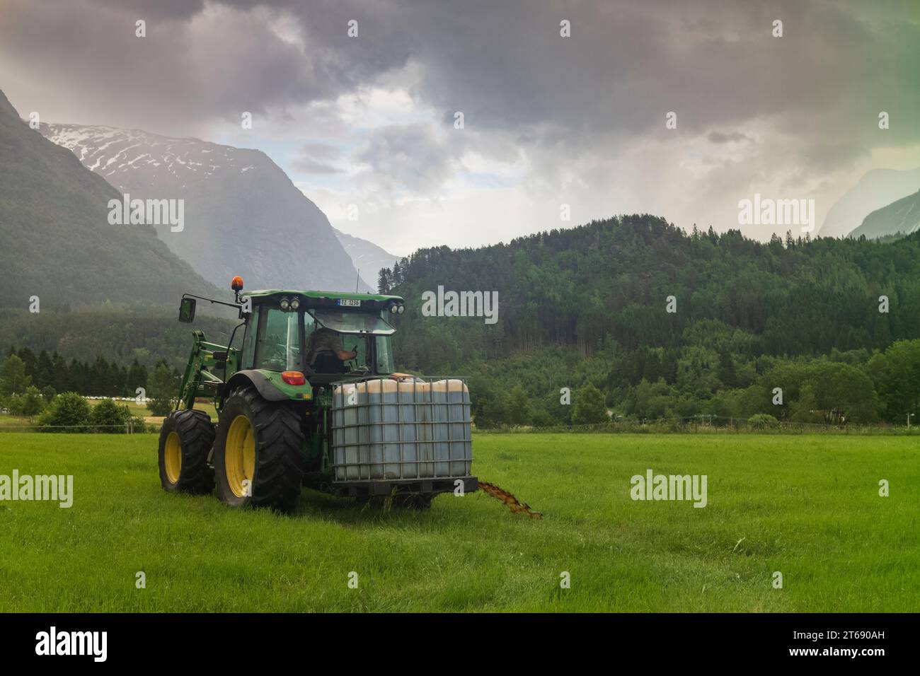 Loen, Norwegen, 26. Juni 2023: Ein Landwirt fährt seinen Traktor auf einem Feld, während er seine Ernte an einem bewölkten Tag düngt. Regen erscheint im Hintergrund. Stockfoto