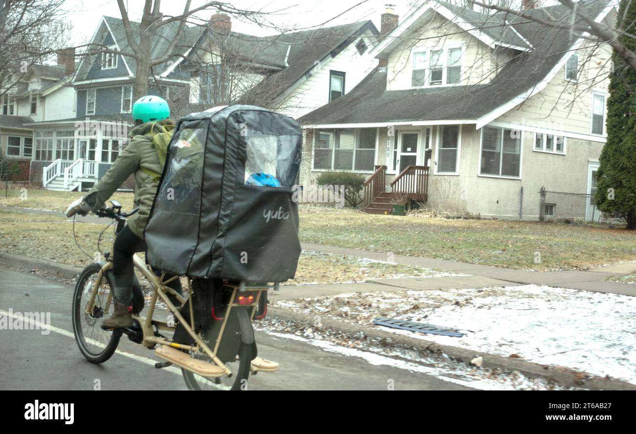 Frau, die im Regen Fahrrad fährt, mit einer schützenden Kindertasche auf dem Rücken und wahrscheinlich einem Kind darin. St. Paul Minnesota MN USA Stockfoto