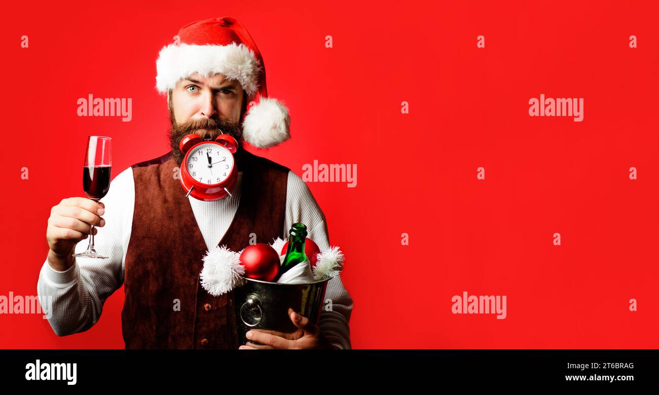 Zeit zum Feiern. Bärtiger Mann mit Weihnachtsmütze mit Wecker. Der Kellner hält ein Glas Wein und einen Eimer mit einer Flasche Champagner und Weihnachtsdekoration Stockfoto