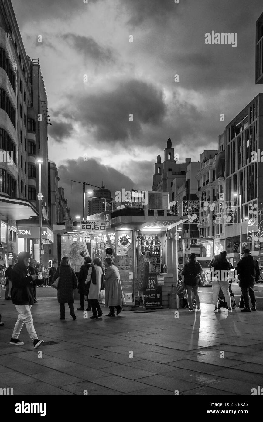 Schwarz-weiß-Stil von Menschen, die in der Nähe eines Kiosks für alkoholfreie Getränke und Süßigkeiten vorbeifahren, mit dunkelgrauen Wolken im Hintergrund in Callao bei Nacht Madrid Spanien Stockfoto