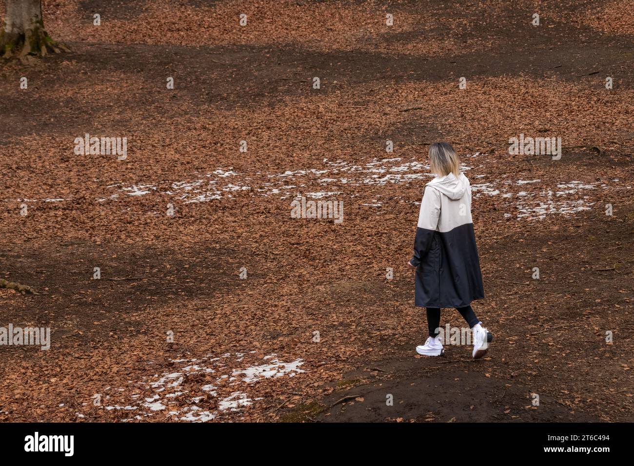In einem langen Mantel und weißen Turnschuhen spazieren durch einen Park mit gefallenen Blättern und Schneeflächen auf dem Boden Stockfoto