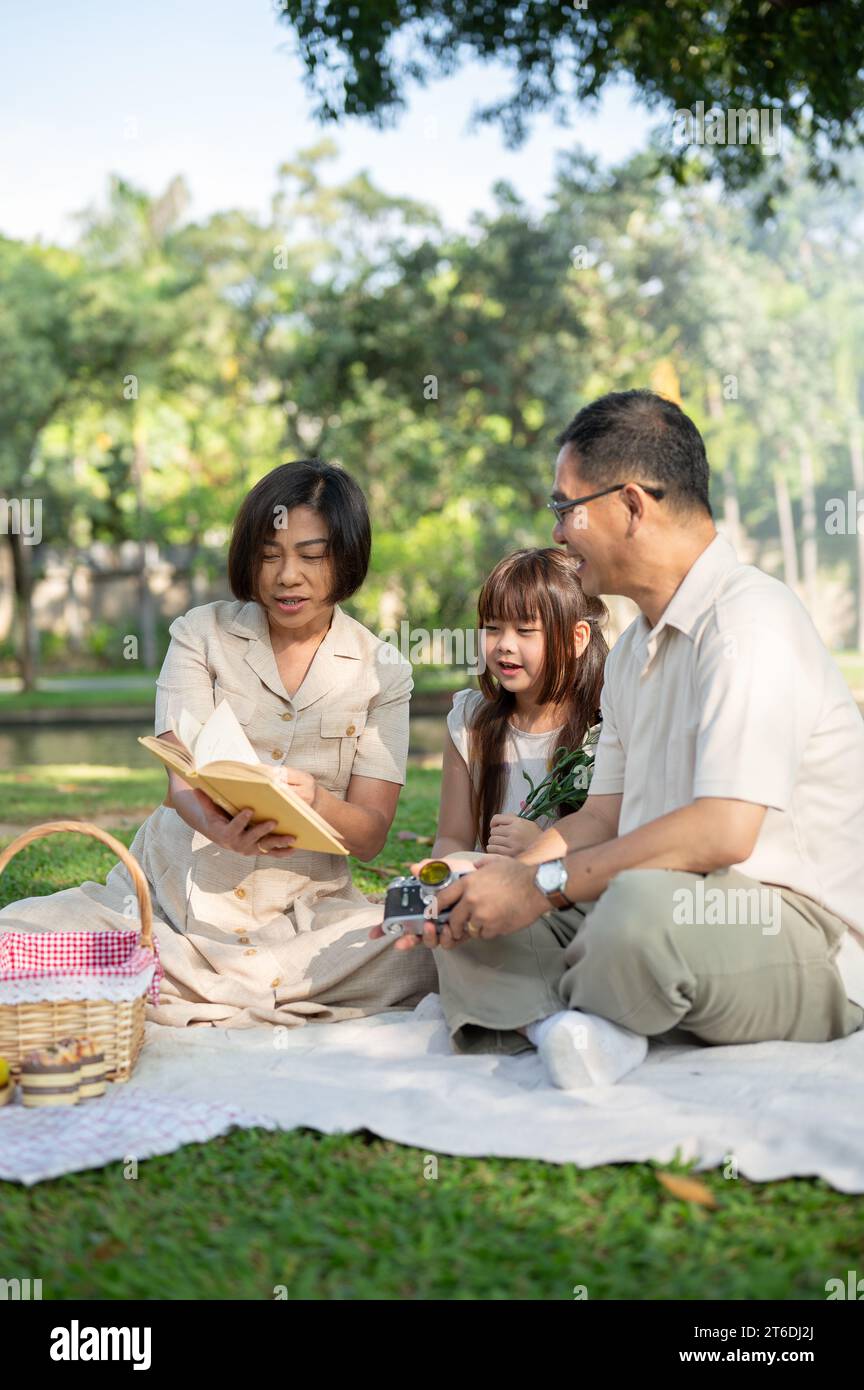 Schöne asiatische Großeltern haben Spaß mit ihrer Enkelin und genießen es, ein Buch zu lesen, während sie am Wochenende gemeinsam in einem Park picknicken. Familie Stockfoto